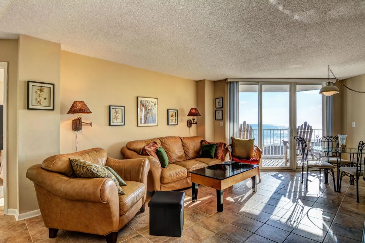A spacious, sunlit living area featuring a leather sofa and coffee table, with large glass doors opening onto a balcony. The view outside reveals the ocean, and the inviting layout includes a dining area, all styled in a modern coastal decor. The soft lighting and comfortable seating create a relaxing and welcoming atmosphere.
