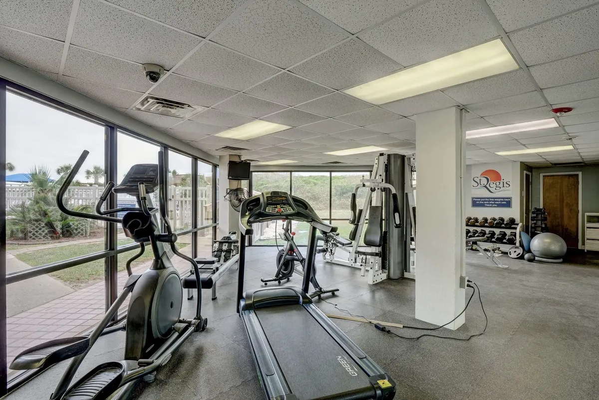 A bright and energetic interior fitness center featuring a treadmill, elliptical machine, weight training equipment, and large windows letting in plenty of natural light. The mood is motivating, with space for both solo workouts and group fitness activities.