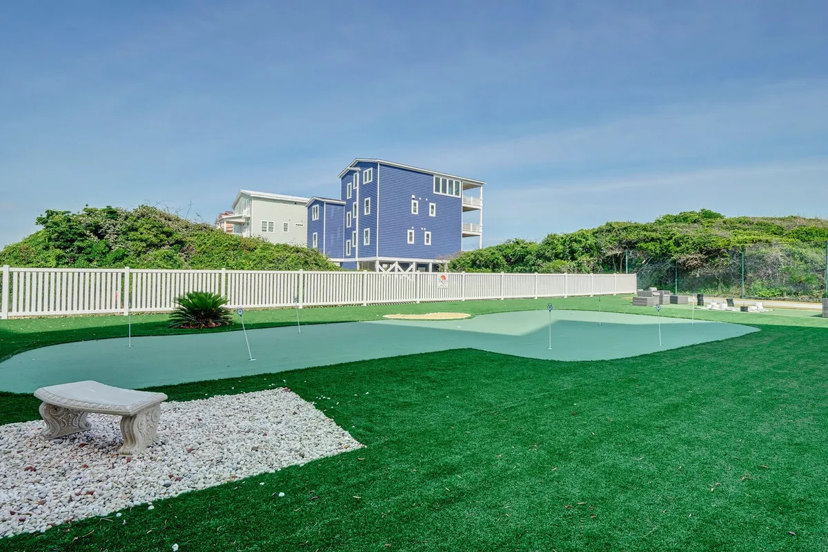 The image captures a vibrant exterior scene featuring an artificial putting green set amidst a beautifully landscaped garden. In the background, a blue and white house adds to the inviting atmosphere, while the garden offers ample space for gathering and relaxation. The foreground encourages outdoor games and social interaction, ideal for group activities.