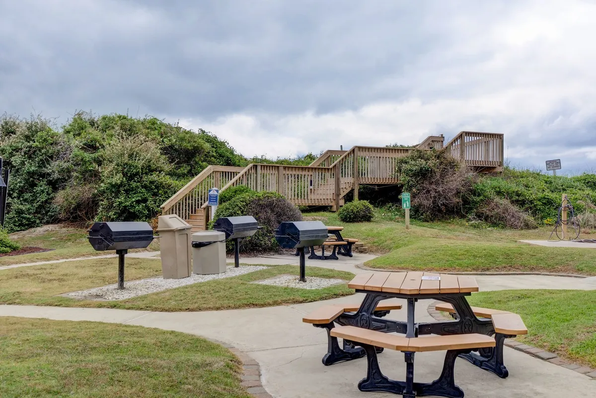An inviting barbecue and picnic area with several picnic tables, barbecue grills, and lush greenery, set beside a staircase. The mood is communal and relaxed—perfect for sunset barbecues or socializing with friends and family outdoors.