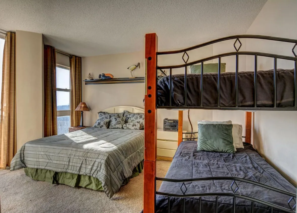 A family-friendly bedroom is shown, including a queen bed and a twin-over-twin bunk bed, as well as shelves with decorative touches and windows dressed in curtains. The atmosphere is warm and playful, suitable for both kids and adults, and well-suited for groups or families.