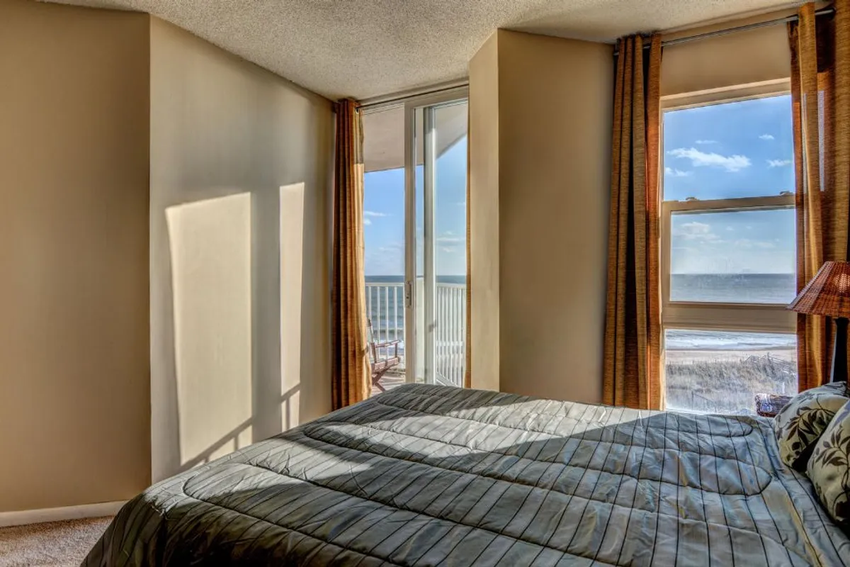 A tranquil bedroom with large windows framing an ocean view, a neatly made bed, and flowing curtains. The serene setting emphasizes opportunities for rest, relaxation, and inspiration, bridging the workday with moments of peaceful retreat.