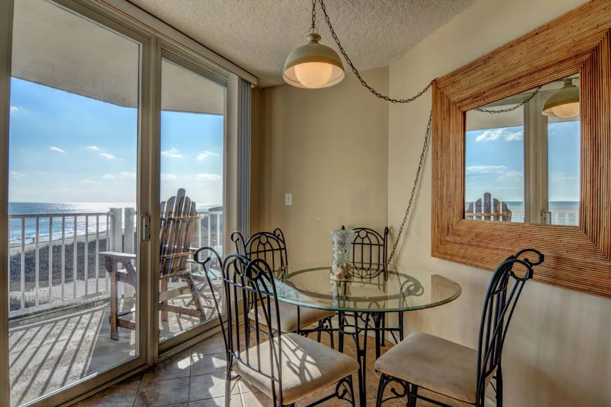 A relaxed dining space featuring a round glass table with metal chairs and large glass doors that open to a balcony with an ocean view. The setting is bathed in natural light, creating an airy, tranquil environment perfect for sharing meals, sipping coffee, or enjoying the fresh ocean breeze.