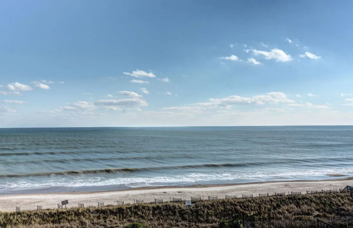 The image showcases a sweeping view of North Topsail Beach, with gentle ocean waves rolling onto a wide sandy shore bordered by natural dune grass under a serene sky with soft clouds. The tranquil scene suggests opportunities for sunrise yoga, evening bonfires, watching dolphins, and relaxing beachside activities.