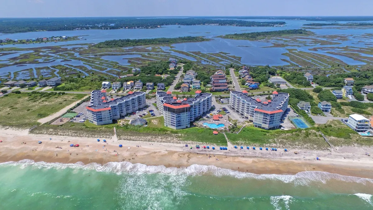 This image captures the inviting beachfront buildings, a vibrant swimming pool, sandy beach, and gentle ocean waves, all under colorful umbrellas. Families are seen swimming, relaxing by the pool, and enjoying playful moments on the beach, embodying classic seaside fun and togetherness.