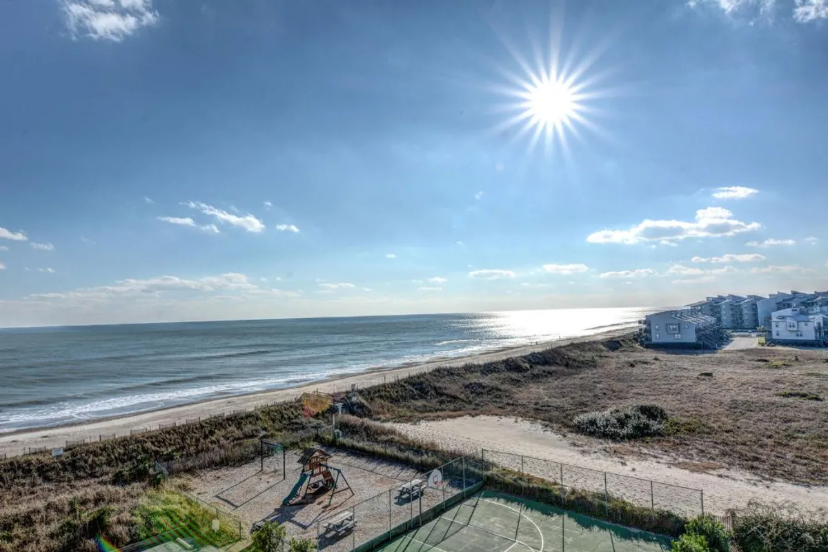 A wide view of the ocean with a playground and basketball court in the foreground. Children gather and play at the playground while others enjoy fun on the court, set against a backdrop of sunlit waves and endless blue sky.