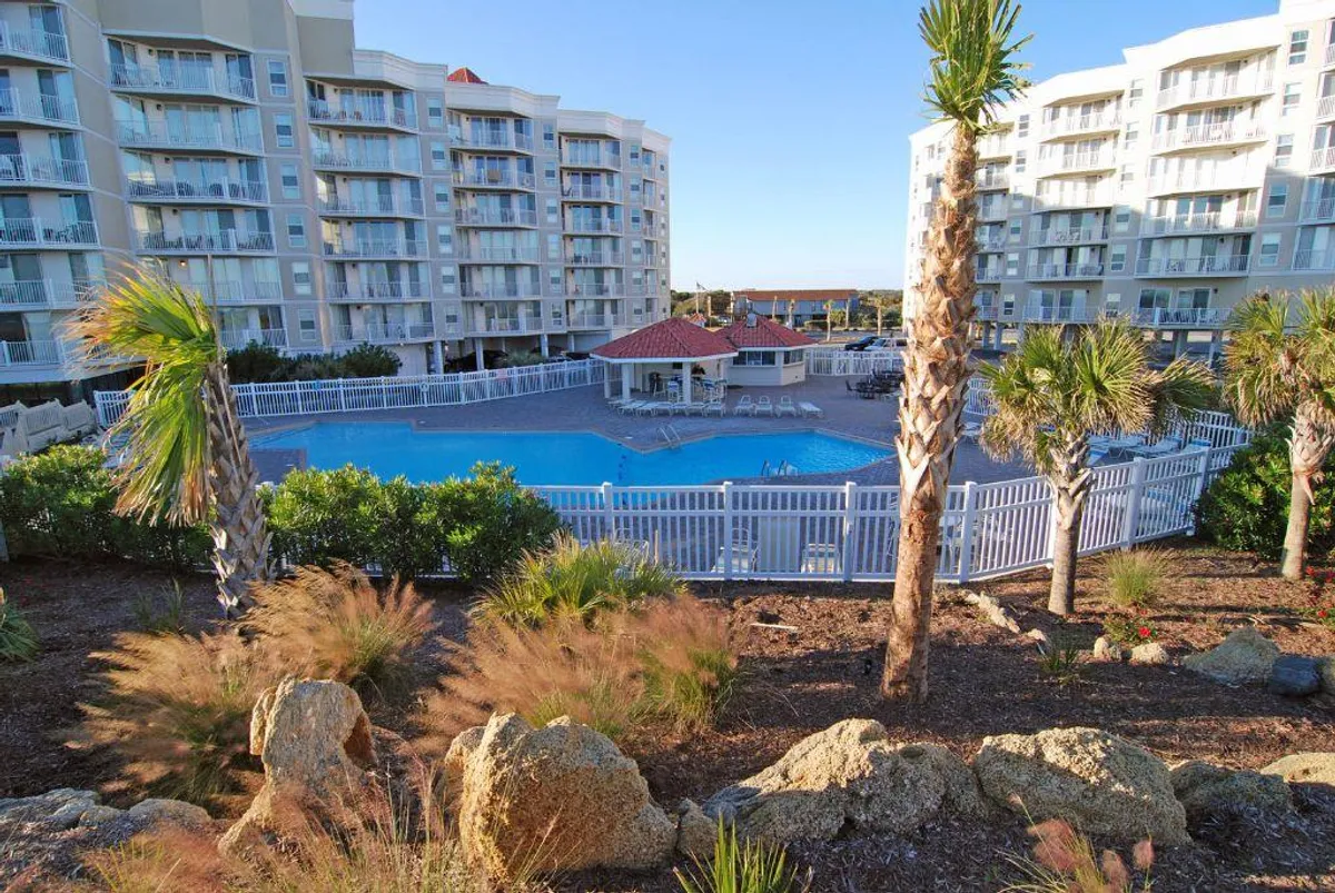 A tranquil and inviting view of the resort's main swimming pool framed by palm trees, with surrounding buildings visible. There are ample poolside spaces for lounging or social gatherings, and the landscaped setting enhances the resort atmosphere.