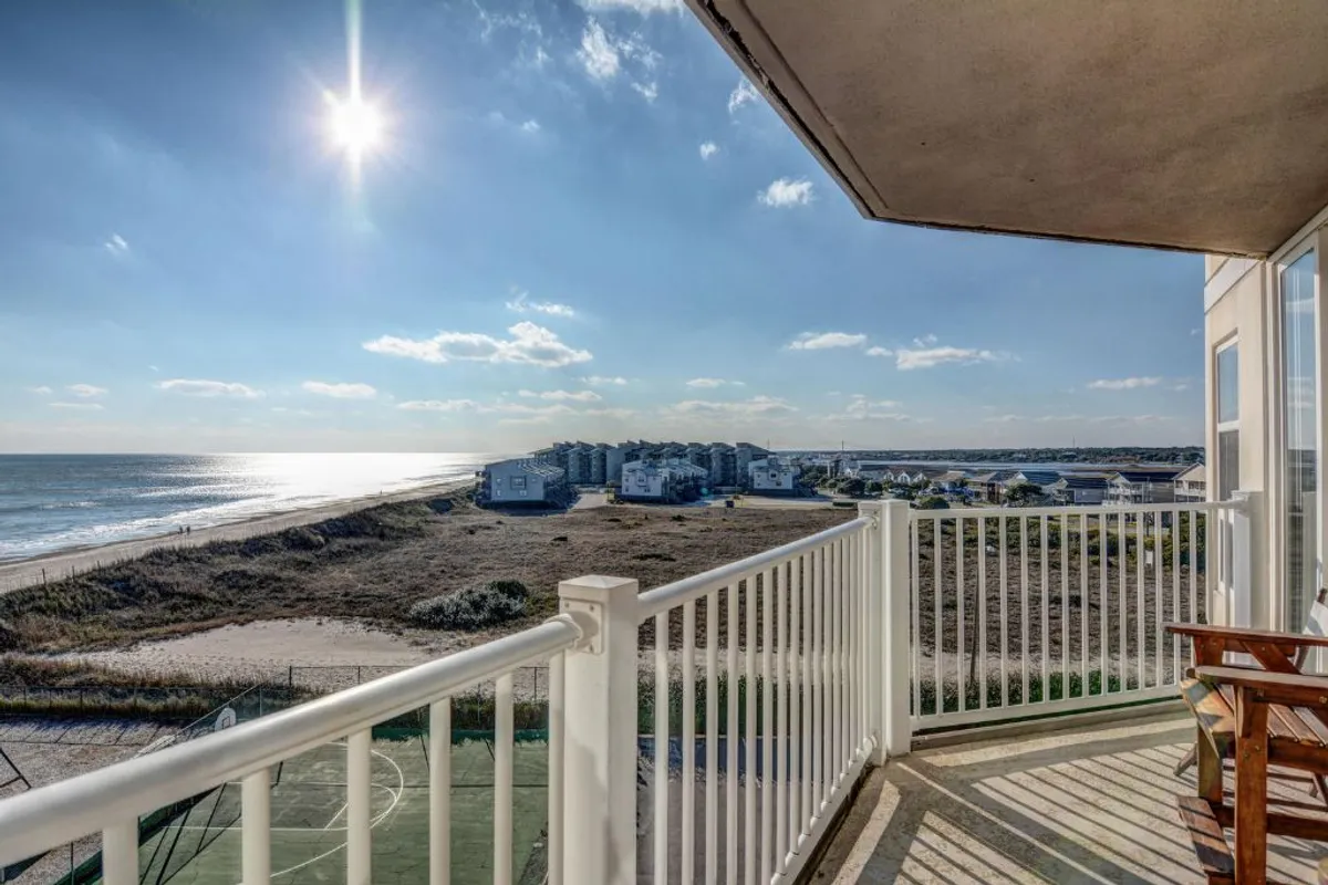 A serene balcony scene with sunlight streaming over the ocean. The balcony railing frames a sweeping view of the beachfront and gentle waves, inviting relaxation and quiet contemplation. The mood is peaceful and the setting ideal for morning coffee or watching the sunset.