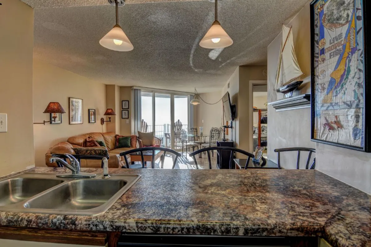 This image showcases a spacious, open-plan kitchen and dining area with a kitchen counter, ample dining space, cozy living room seating, and large windows offering a glimpse of the outdoor view. The overall vibe is warm, inviting, and conducive to group gatherings or intimate family meals.