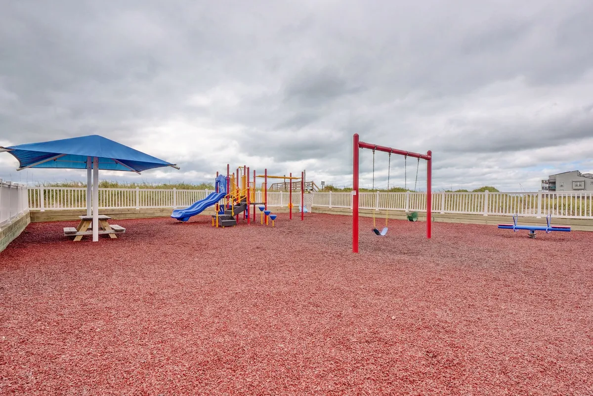 A cheerful exterior shot of a modern playground area with swings, colorful play equipment, and a picnic area with a safe surface beneath. Families and children can be seen enjoying the space, creating a joyful and welcoming atmosphere.